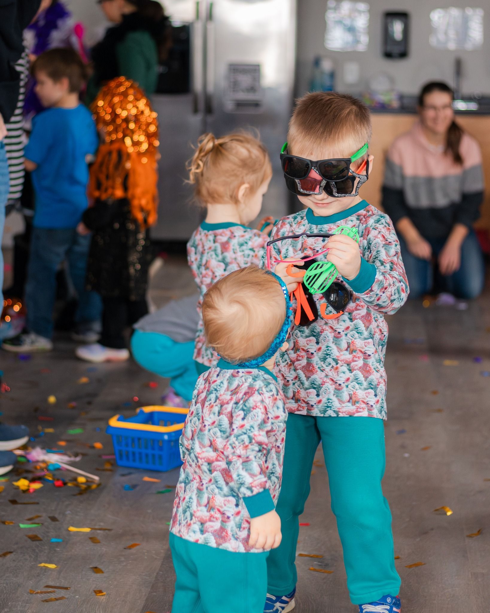 Three young children play indoors; one wears a mask and holds a toy, another looks on.
