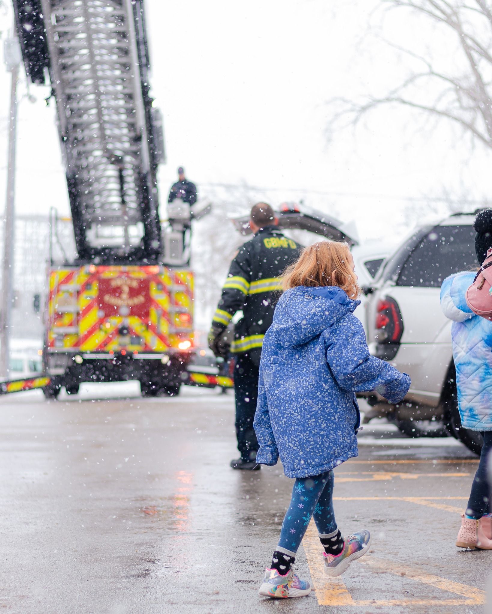 Fire truck with raised ladder, firefighters, and children walking during snowfall.