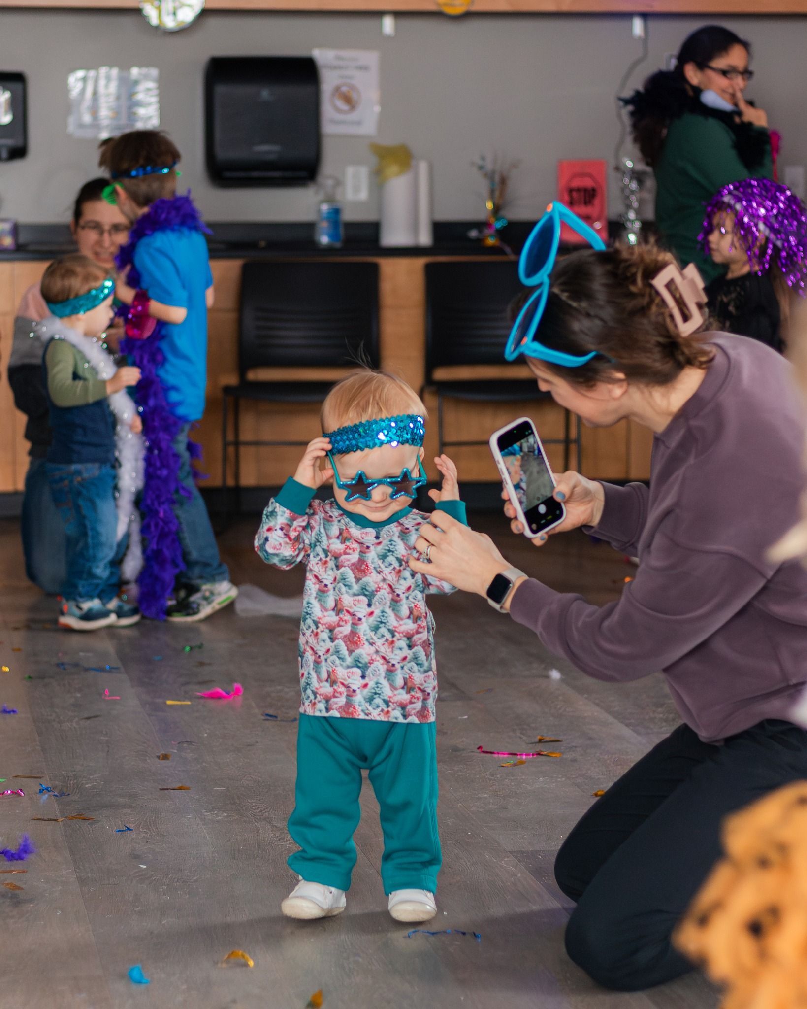 Woman taking photo of child in party attire; others in background with party accessories.