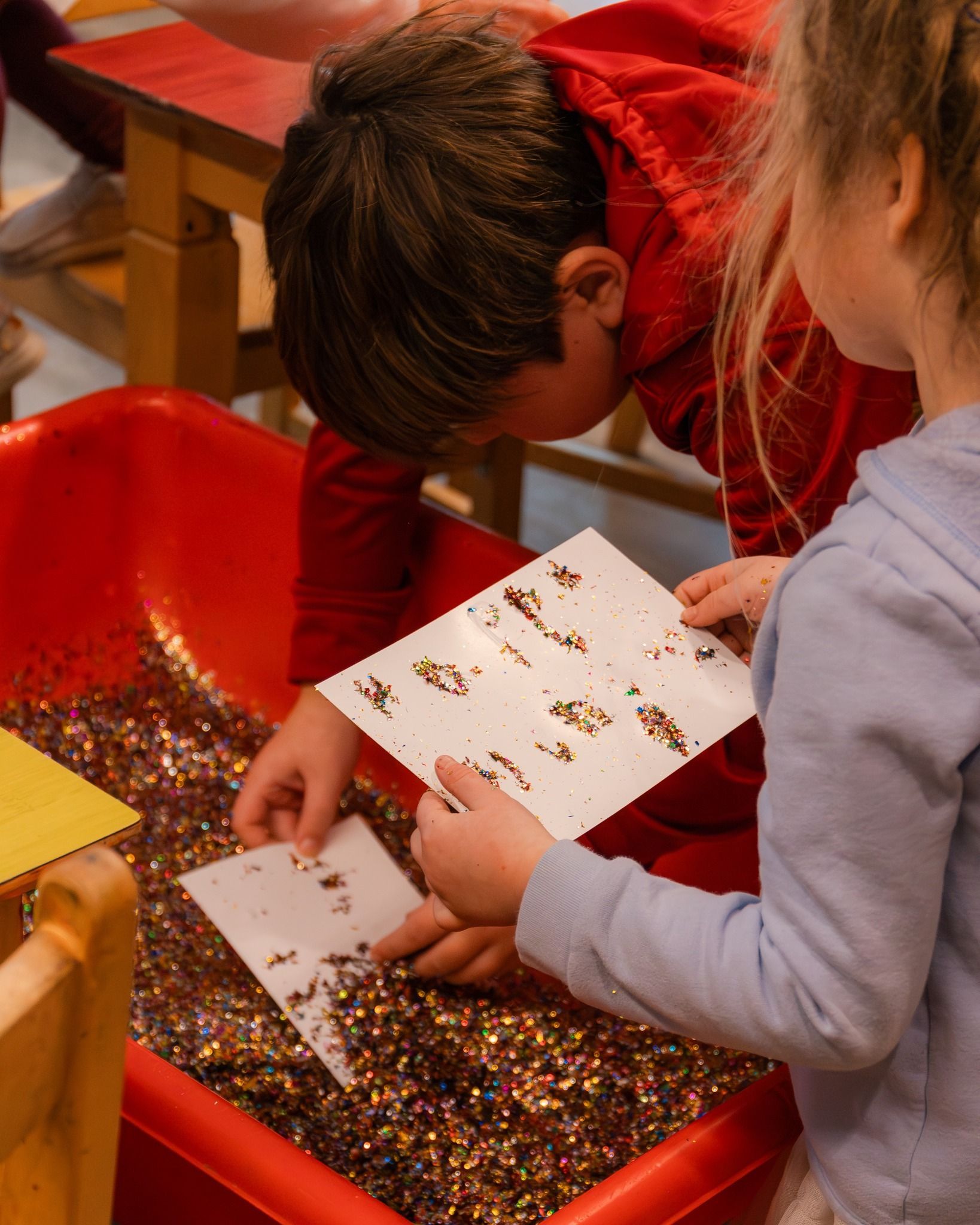 Children examining a sheet of paper with small objects in a red bin.