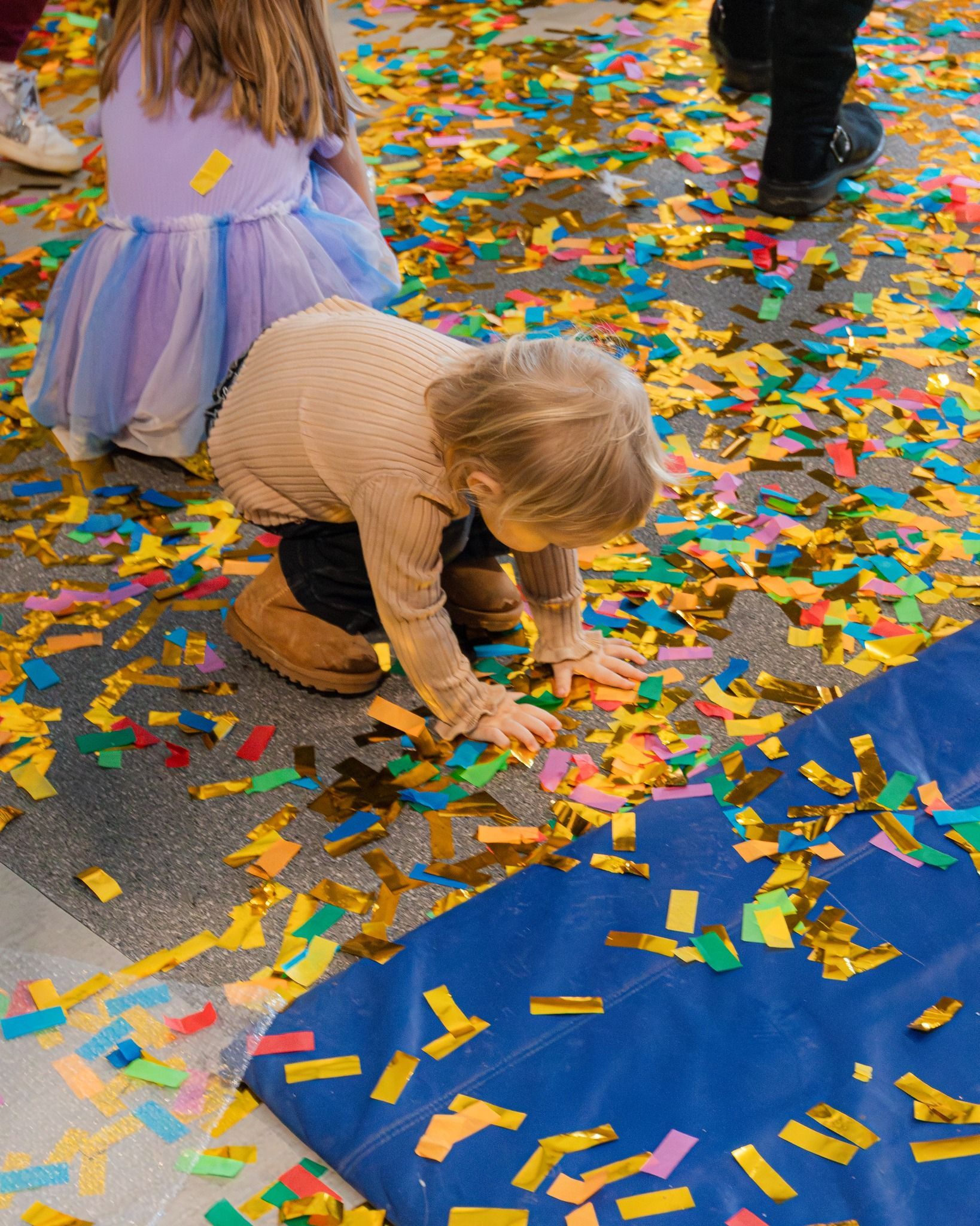 Child playing in confetti on the floor; another child in a tutu is nearby.