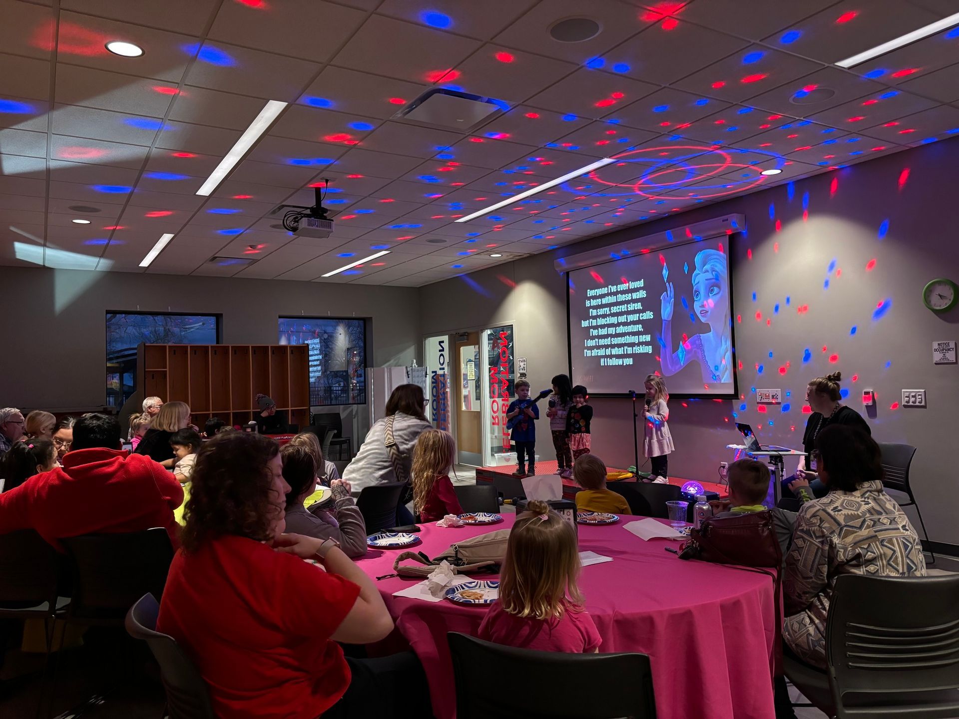 Event in a room with pink tables, stage with children performing, and red/blue lights on the ceiling.