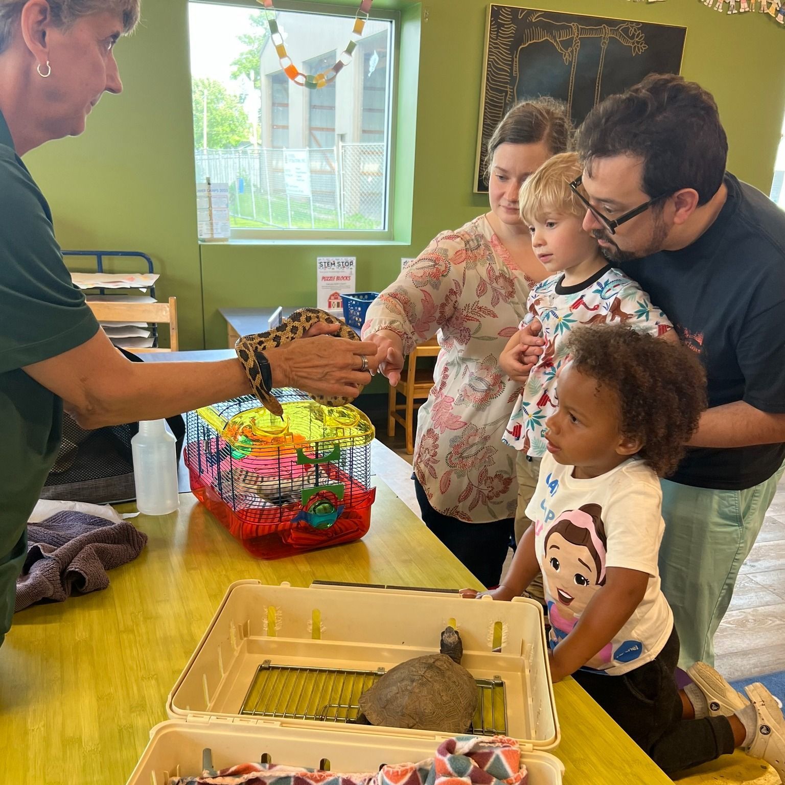 A woman shows a family a leopard gecko in a classroom. Children look on, fascinated.