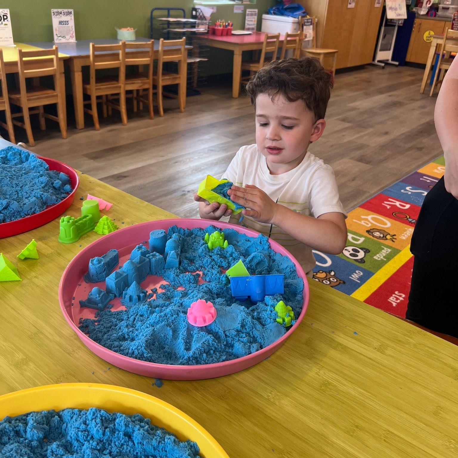 A young boy playing with blue kinetic sand in a pink tray, smiling. Other trays and tables are visible.