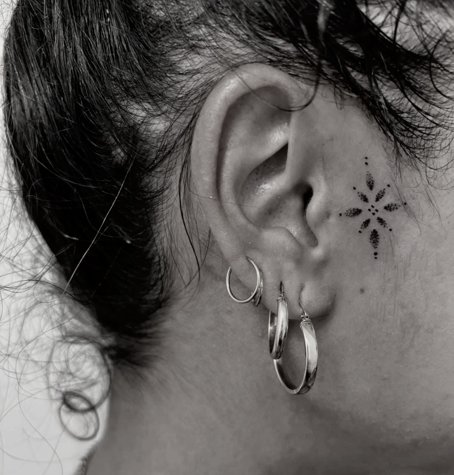 Ear with starburst tattoo, silver hoop earrings, and curly hair pulled back. — Pins and Needles Tattoo in Smithfield, QLD