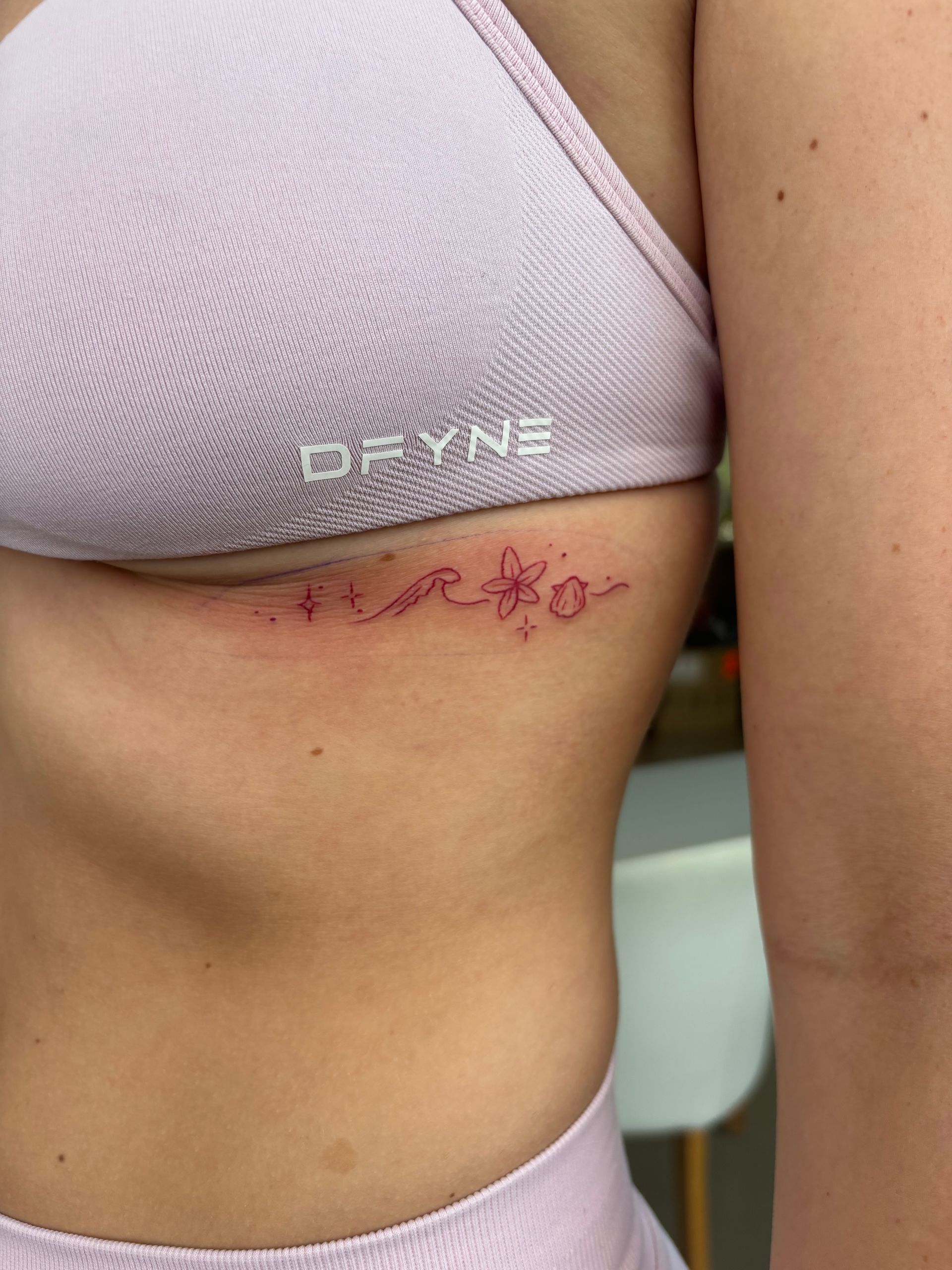 Woman with lavender sports bra, showing side with a thin, purple floral tattoo on her ribs. — Pins and Needles Tattoo in Smithfield, QLD