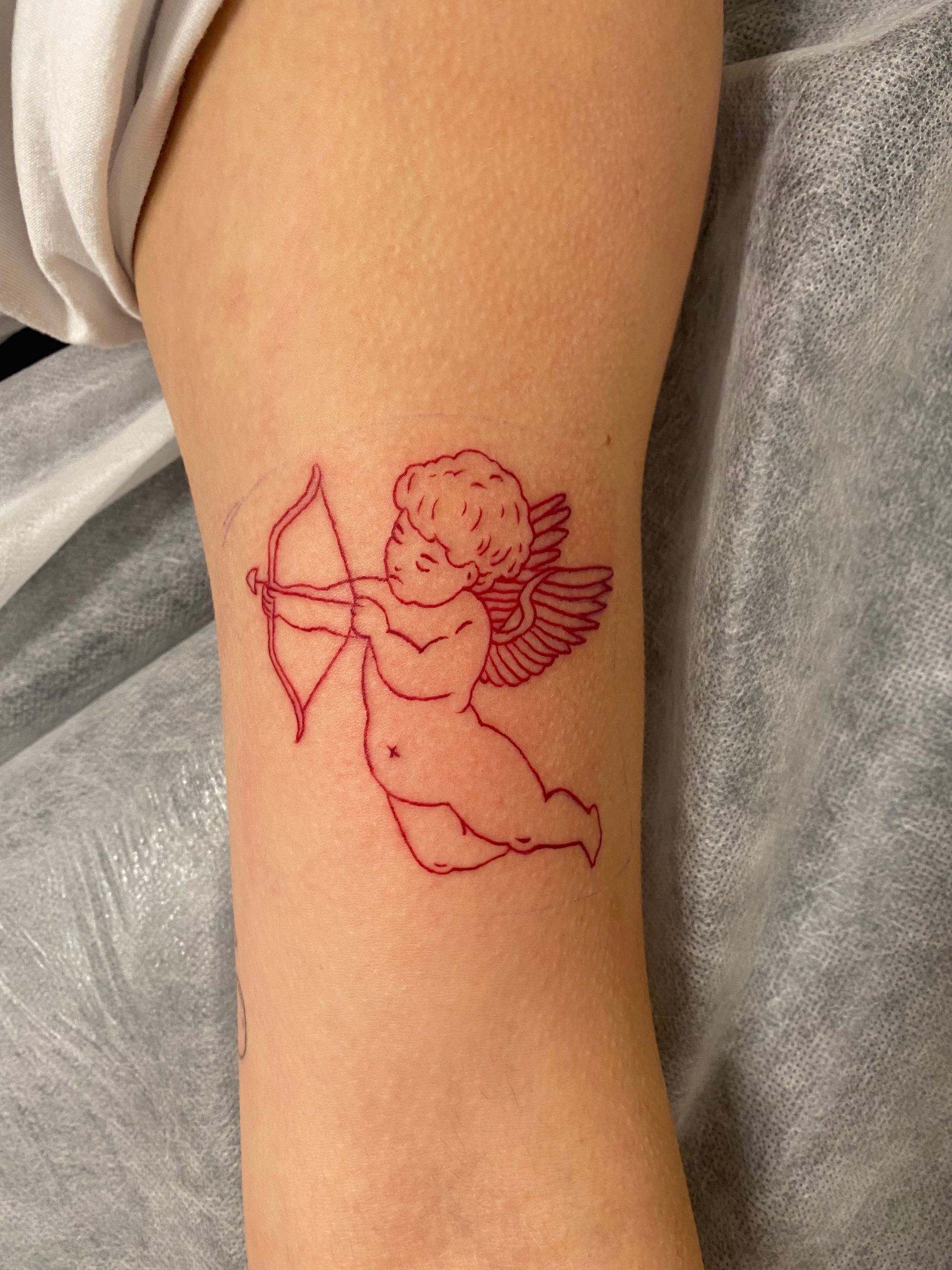 Red outline tattoo of Cupid with bow and arrow on skin. — Pins and Needles Tattoo in Smithfield, QLD