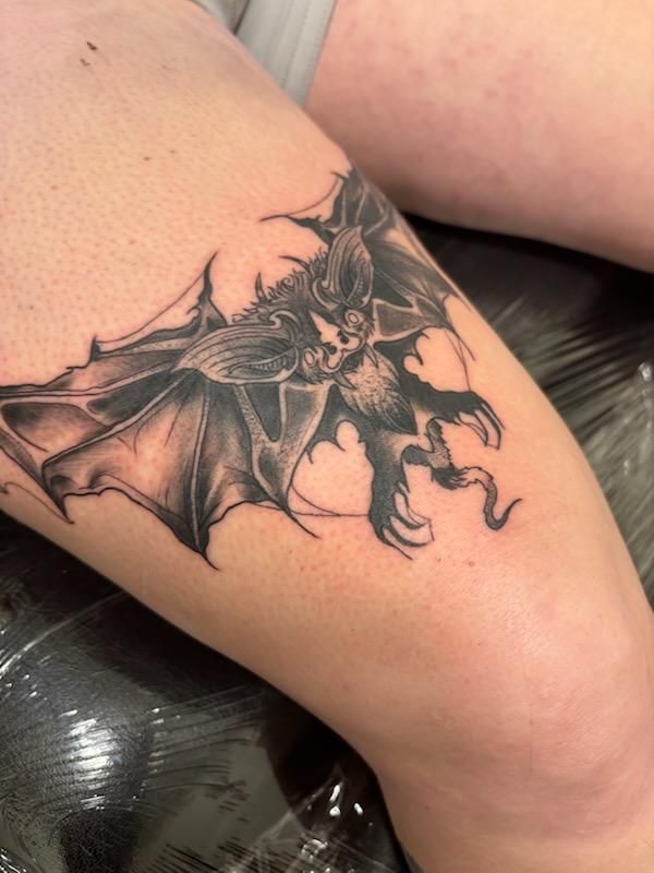 Black and gray tattoo of a bat-winged figure with clawed hands and a skull-like face on a person's leg.