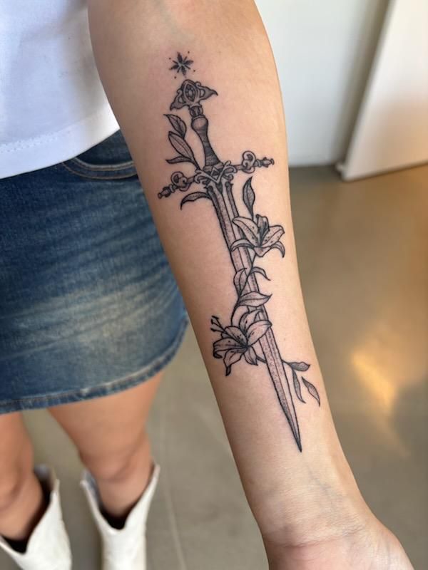 Tattoo of a sword entwined with flowers on a forearm, with a star detail at the top.