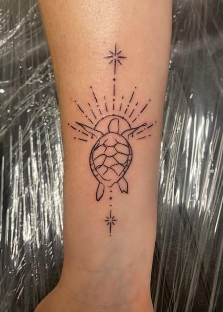 Tattoo of a turtle with sunburst rays and stars on a wrist. Black ink on fair skin.