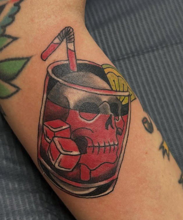 Tattoo of a skull in a drink glass with ice cubes, a straw, and lemon slice, on an arm.