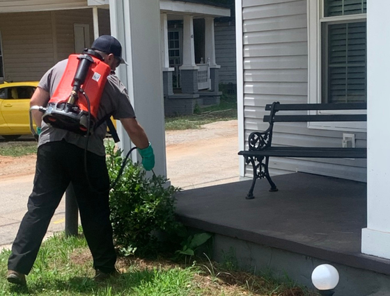Man in black pants and backpack sprayer sprays bush beside a porch.