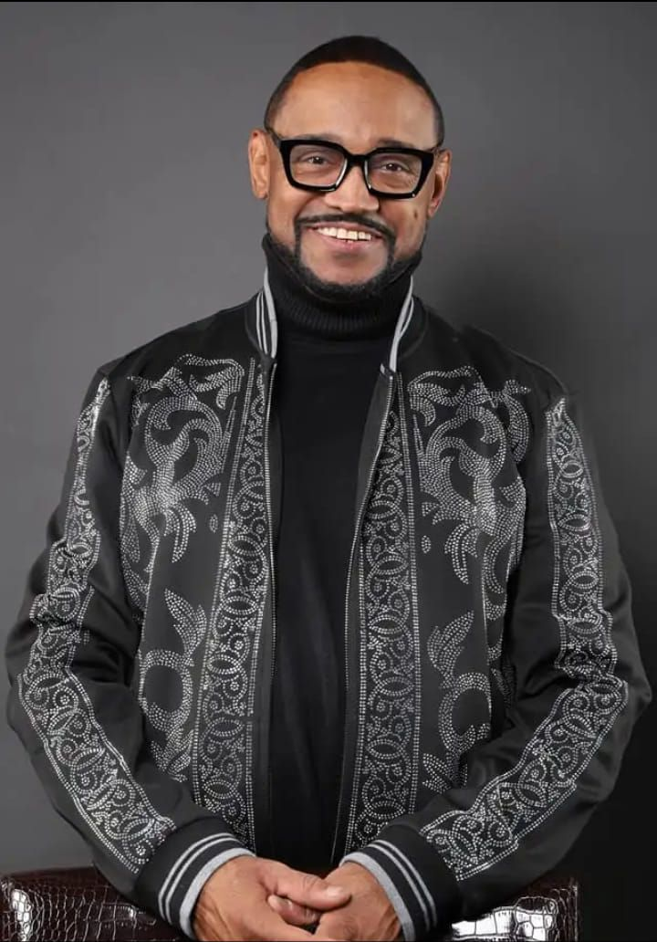 Man in glasses, black turtleneck, and patterned jacket smiles, leaning slightly. Dark background.
