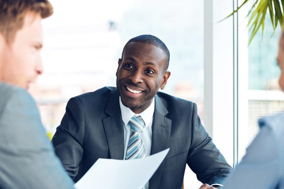 Smiling Black man in a suit at a table with colleagues, holding a paper, in an office setting.