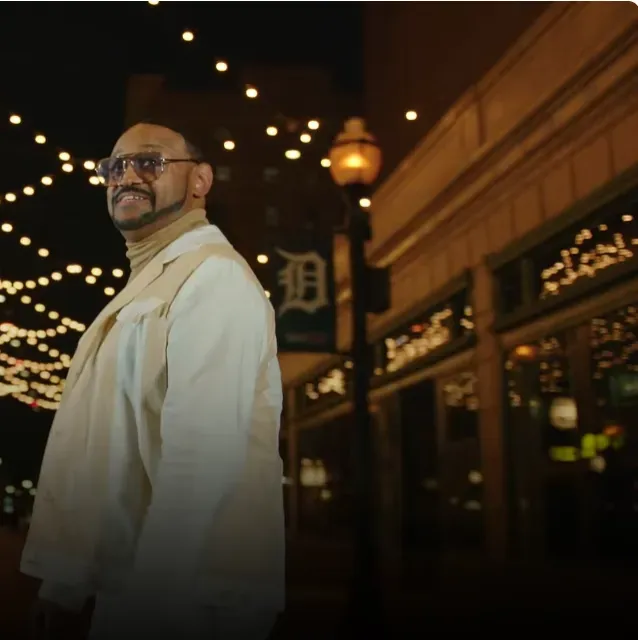 Man in white suit, turtleneck, and glasses, smiling, standing outside building with