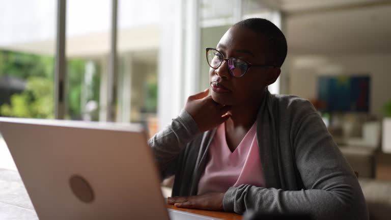 Black woman with glasses, looking thoughtfully at a laptop. She's inside, leaning on her hand.