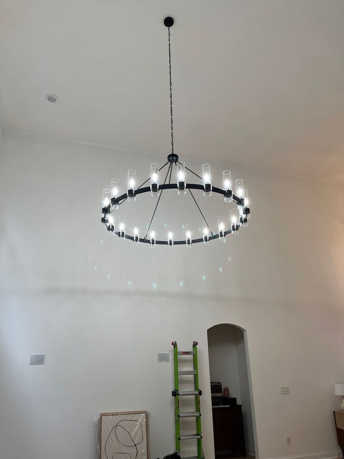 Black chandelier hangs from high ceiling in an empty, white-walled room; green ladder near an arched doorway.