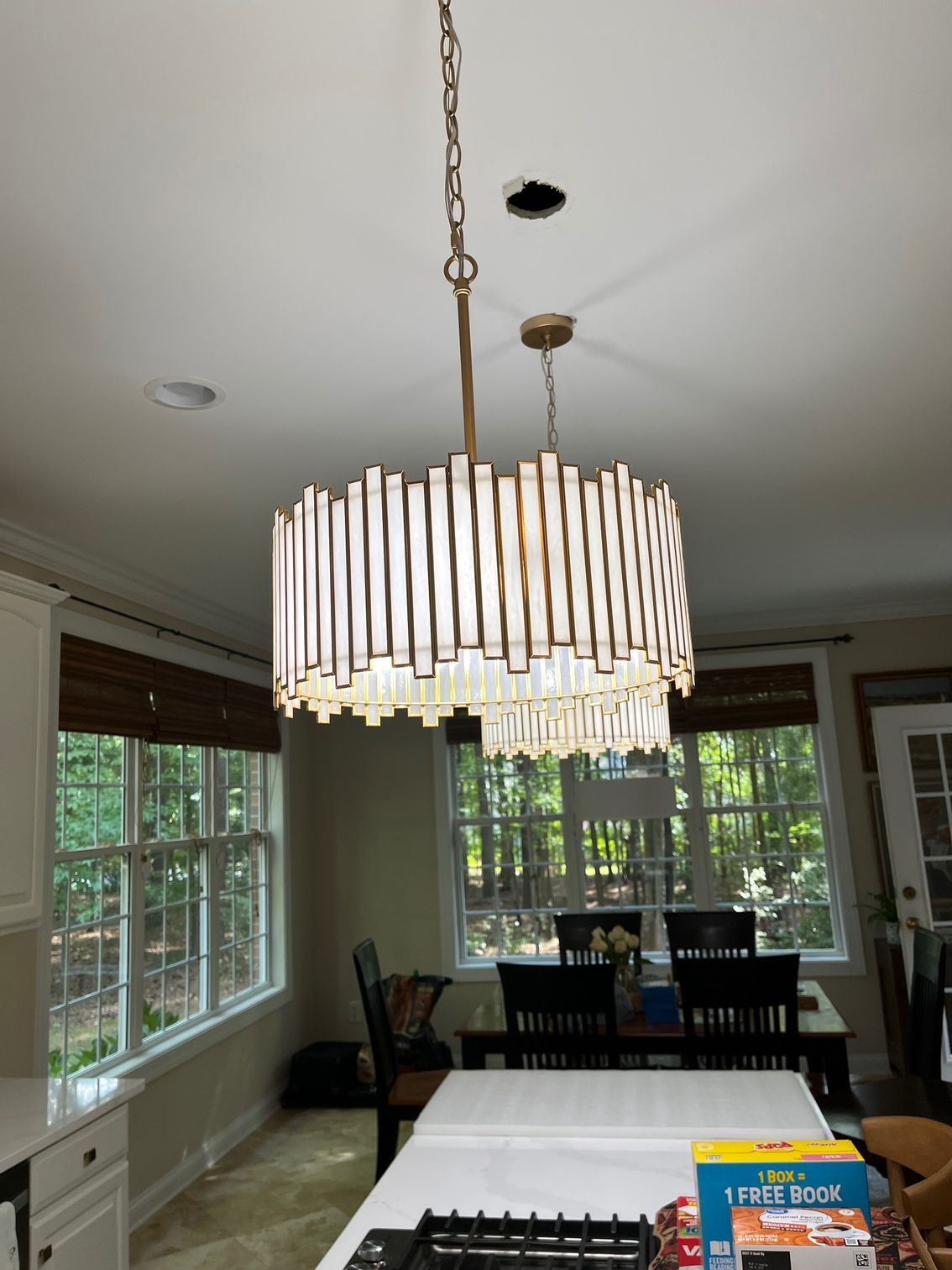 Gold chandelier hangs over a kitchen island with white countertop. Windows show outside trees.