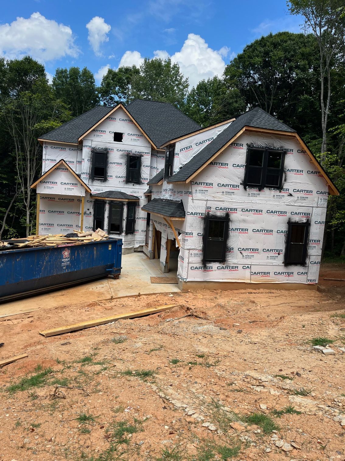 House under construction, covered in white wrap, surrounded by dirt and trees, with a blue dumpster.