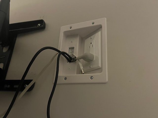 White recessed wall plate with electrical outlet, black cords and white plug. Mounted TV bracket visible on the left.