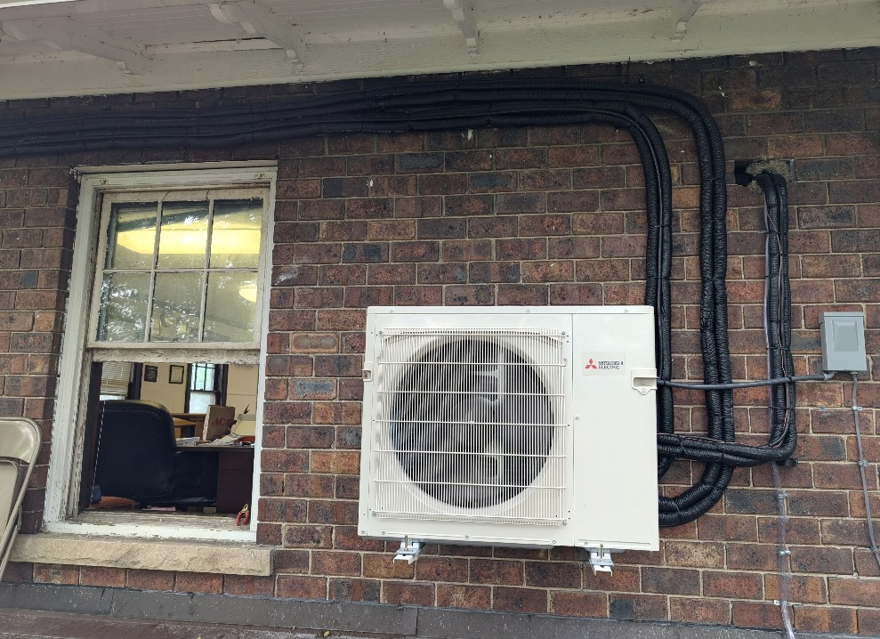 An outdoor HVAC condenser unit mounted on a brick wall next to an open window with insulated piping routed along the wall.