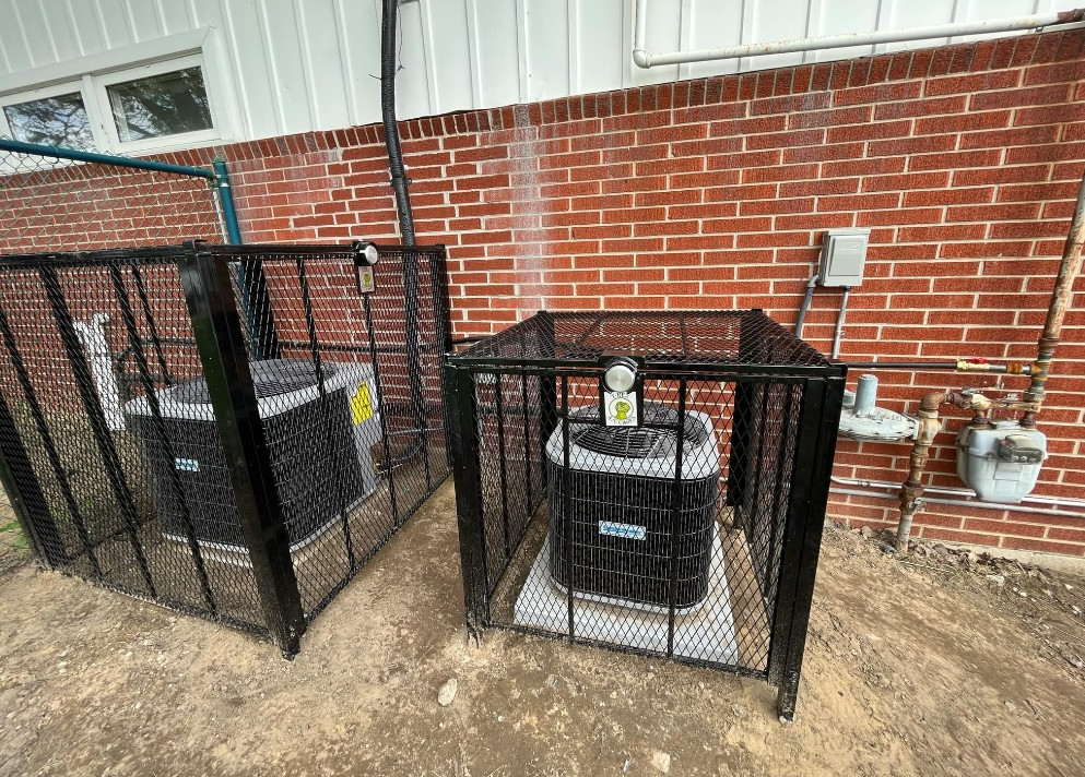 Two black metal security cages protecting HVAC condenser units installed against a red brick exterior wall.