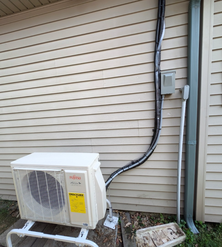 An outdoor mini-split AC unit on a stand against beige siding, with electrical and refrigerant lines running up the wall.