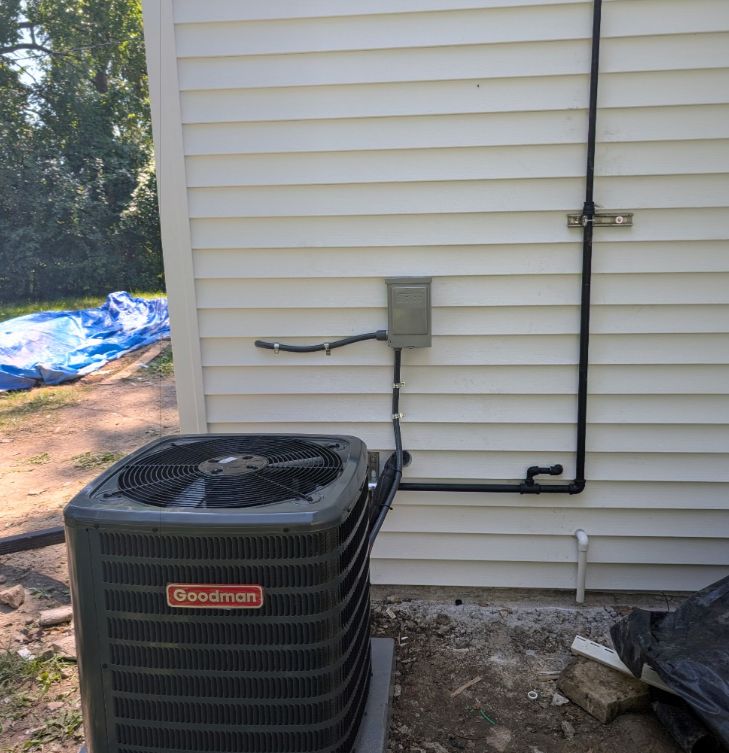 A Goodman outdoor AC condenser unit sits next to a house wall with external electrical and plumbing conduits.