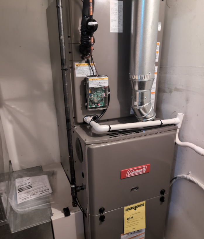 A grey Coleman gas furnace stands vertically in a corner, featuring metal ducting, white PVC pipes, and a control panel.