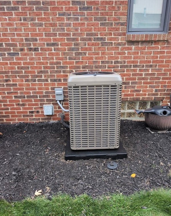 An outdoor HVAC unit sits on a black pad against a red brick wall, next to an electrical disconnect box.