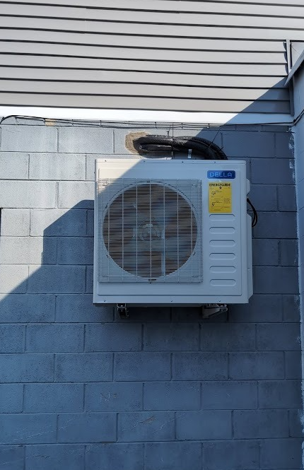 A white mini-split air conditioning unit mounted on a grey cinder block wall under a beige vinyl-sided building.
