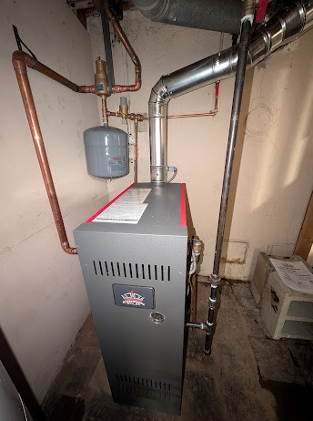 A gray home heating boiler with connected copper plumbing and a silver exhaust vent in a residential basement.