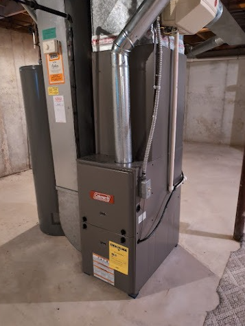 A gray Coleman furnace unit stands in a basement next to a water heater and ductwork.