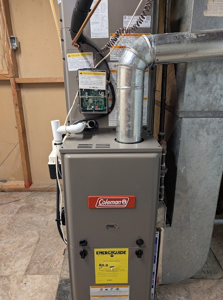 A beige Coleman furnace stands in a utility room with metal ductwork and a white condensate pump attached.