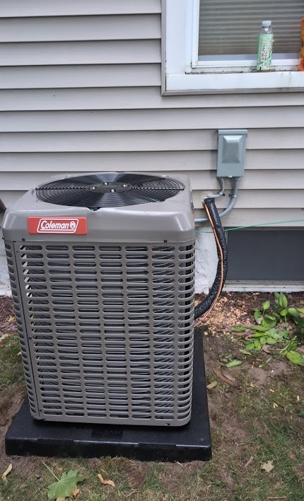 A grey Coleman air conditioning unit sits on a black pad next to a house with beige siding and an electrical disconnect box.