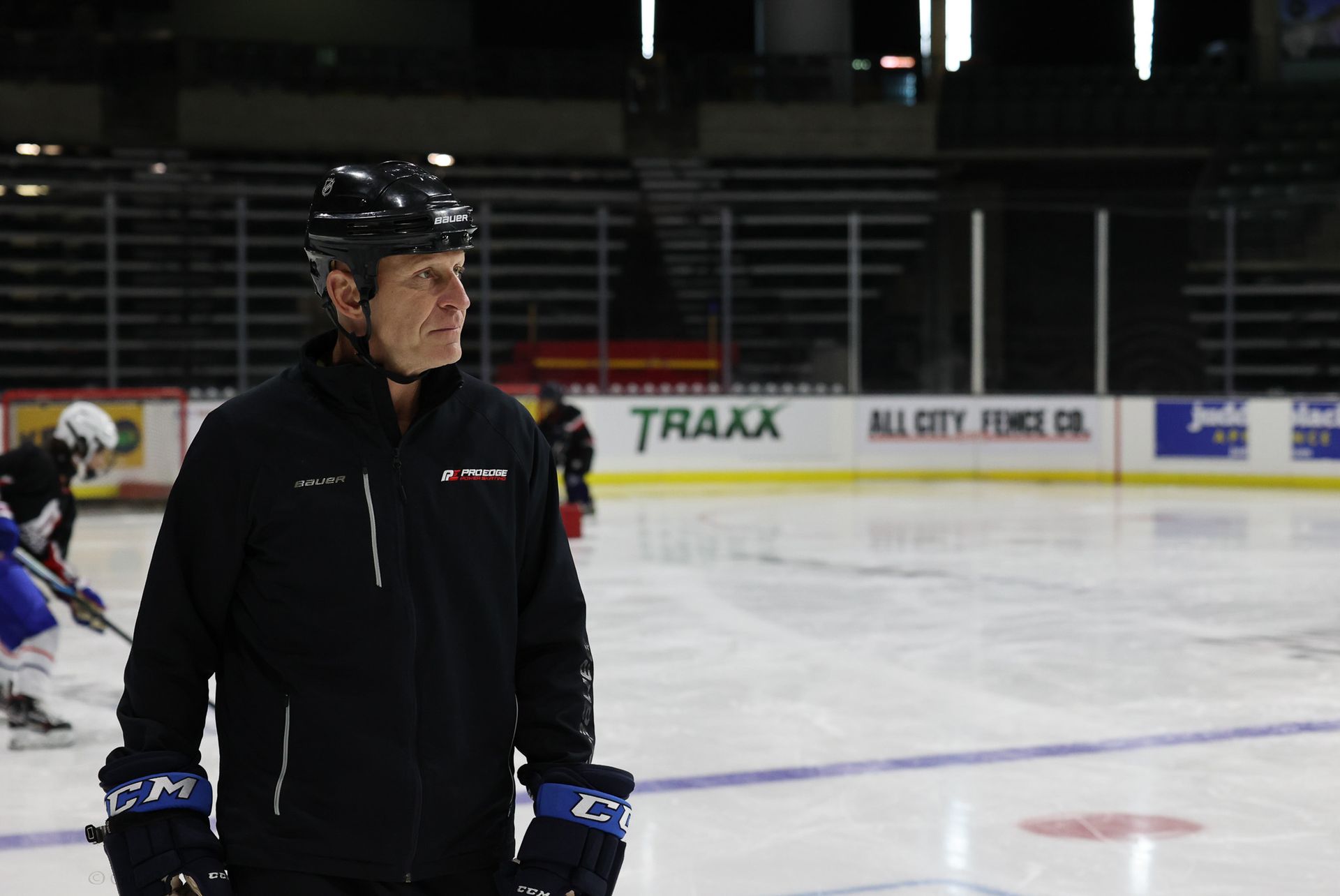 A man in a black jacket is standing on a hockey rink.