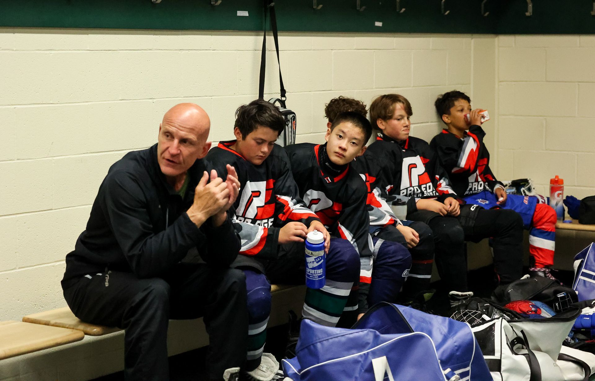 A group of hockey players are sitting in a locker room with a man talking to them