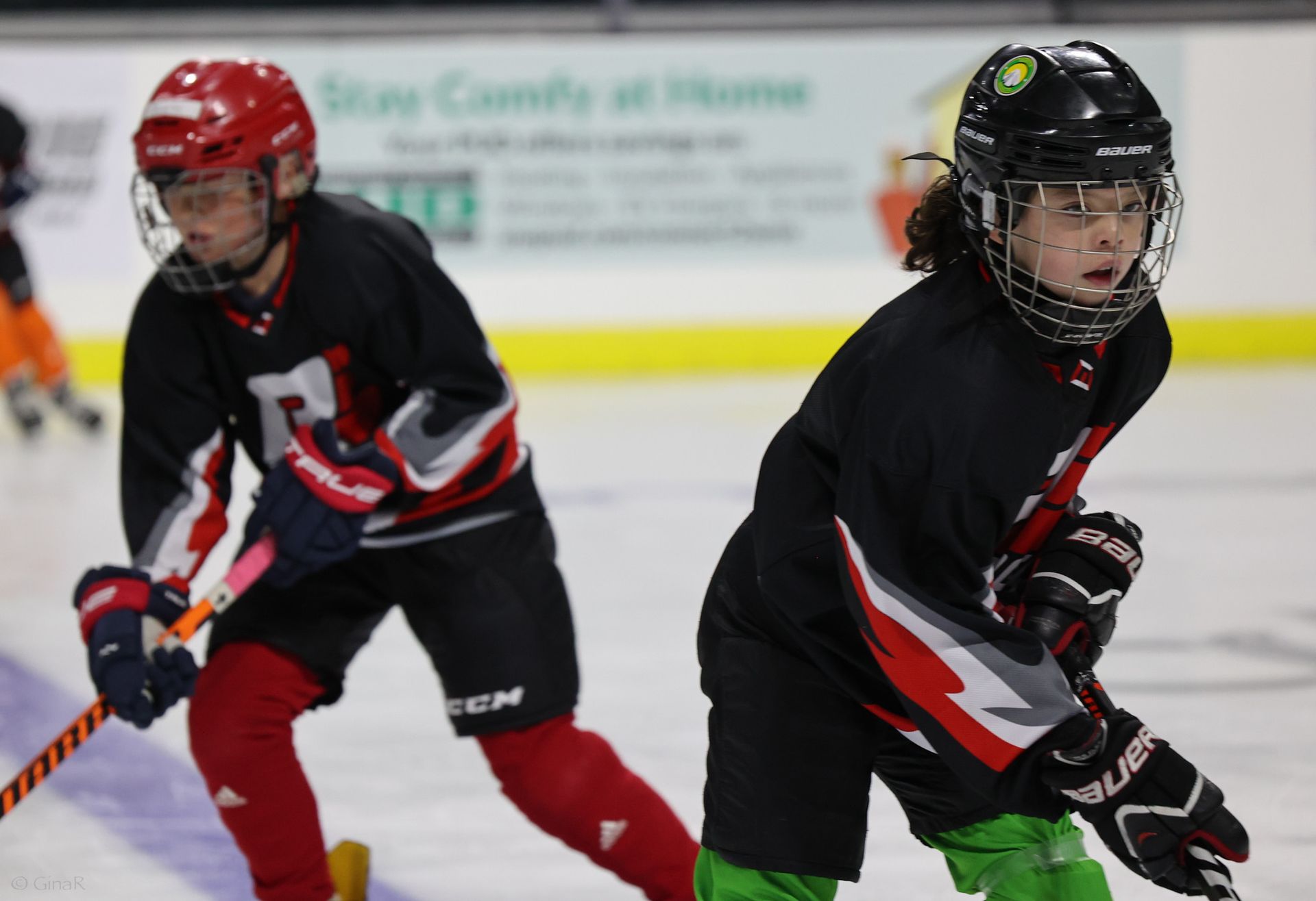 Two boys are playing ice hockey on a rink with a sign in the background that says stay comfy at home