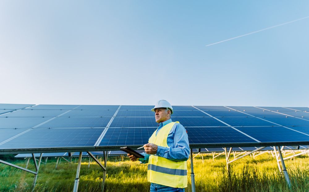 A man is standing in front of a field of solar panels holding a tablet.