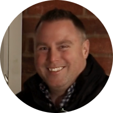 Smiling man with short dark hair, wearing a dark jacket over a plaid shirt, standing in front of a brick wall.