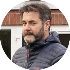 Man with a beard in a blue jacket, standing outdoors in front of a brick building.