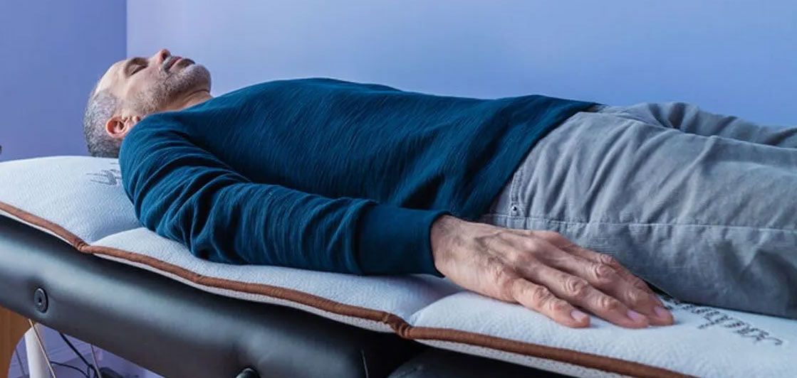 A man in a blue sweater is lying on a massage table with a PEMF mat