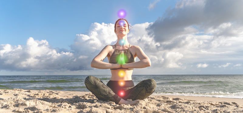Woman meditating on a beach, chakra symbols glowing. Blue sky and sea in the background.