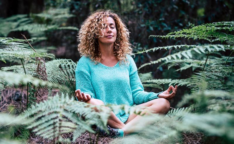 A woman is sitting in a lotus position in the woods surrounded by ferns Grounding with Nature for He