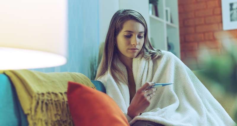 Woman wrapped in a blanket on a sofa, looking at a thermometer. Interior with a lamp, pillows, and a bookcase.