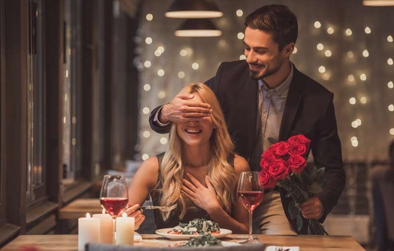 Man surprises woman at dinner with roses, covering her eyes. They are in a dimly lit restaurant.