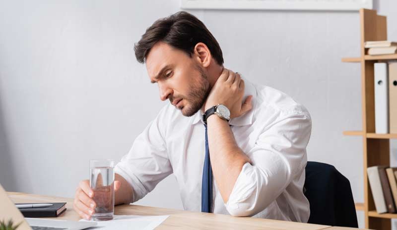 Man at desk with neck pain, holding a glass of water, looking down in an office setting.
