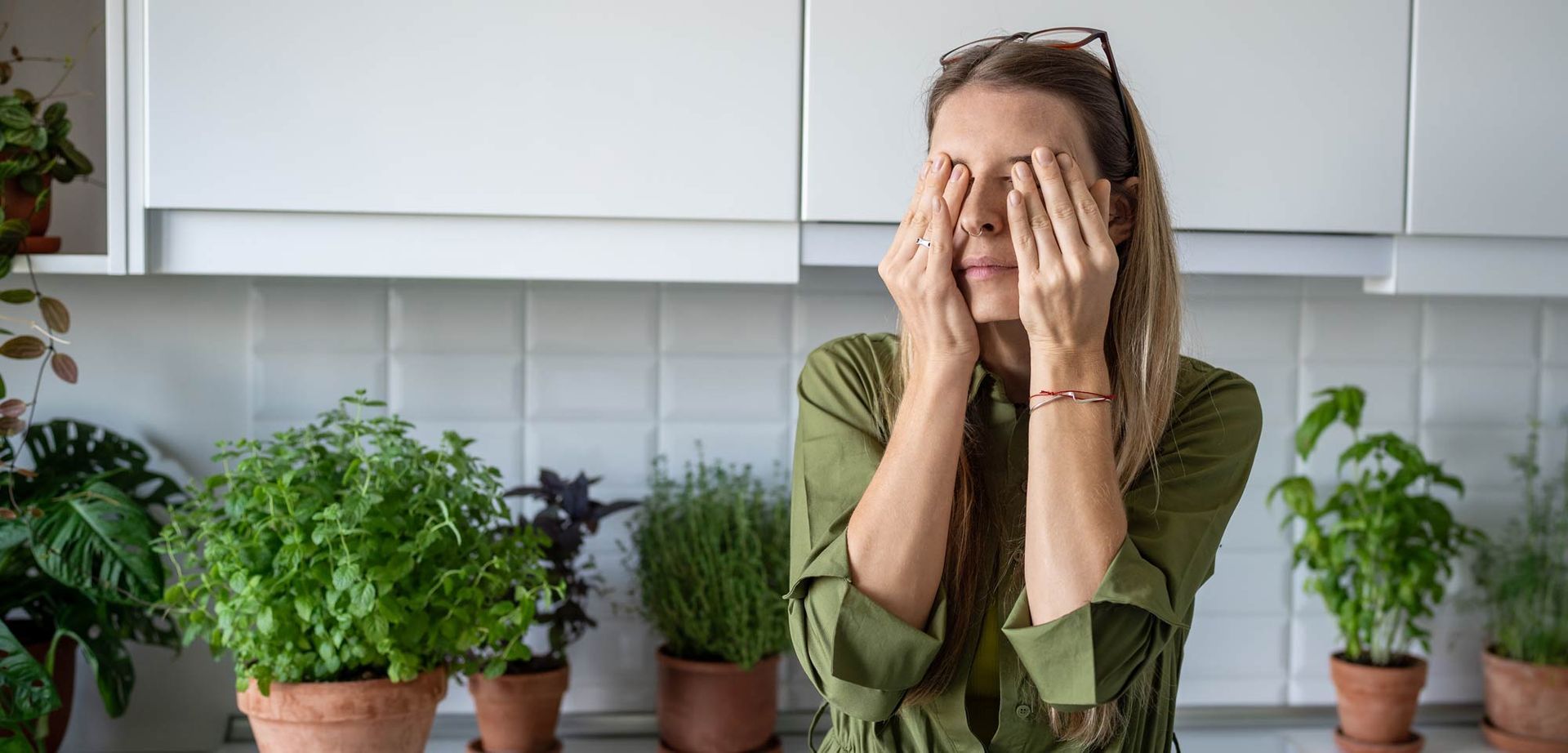 A woman with PDMM is covering her eyes with her hands in a kitchen.