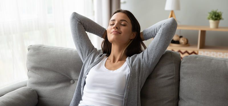 Woman relaxing on a couch, arms behind head, eyes closed. Indoor setting, natural light.