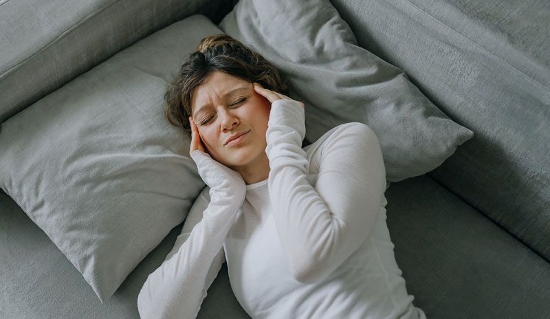 Woman lying on a gray couch, holding her temples, looking distressed. Root-Cause Care
